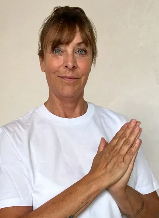 Expert en masaje de Pluma con camiseta blanca, juntando las manos frente al pecho y sonriendo amablemente.