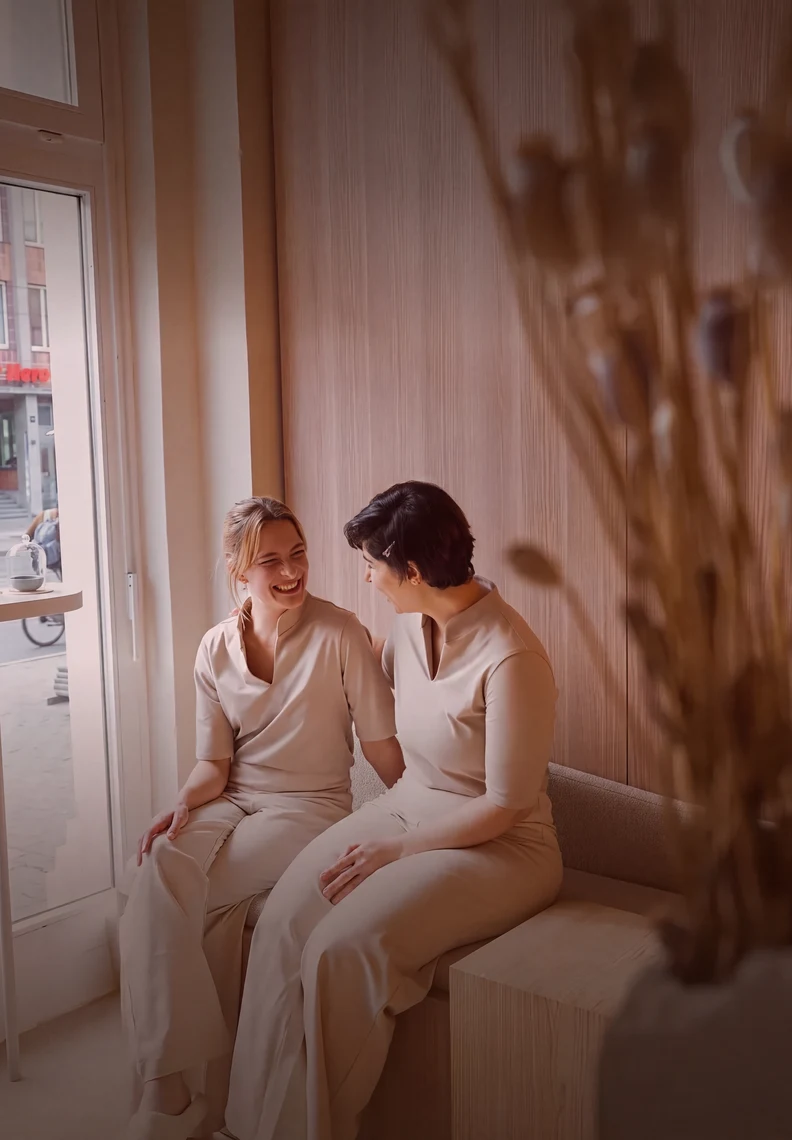 Dos mujeres con ropa beis a juego se sonríen sentadas junto a una ventana luminosa.