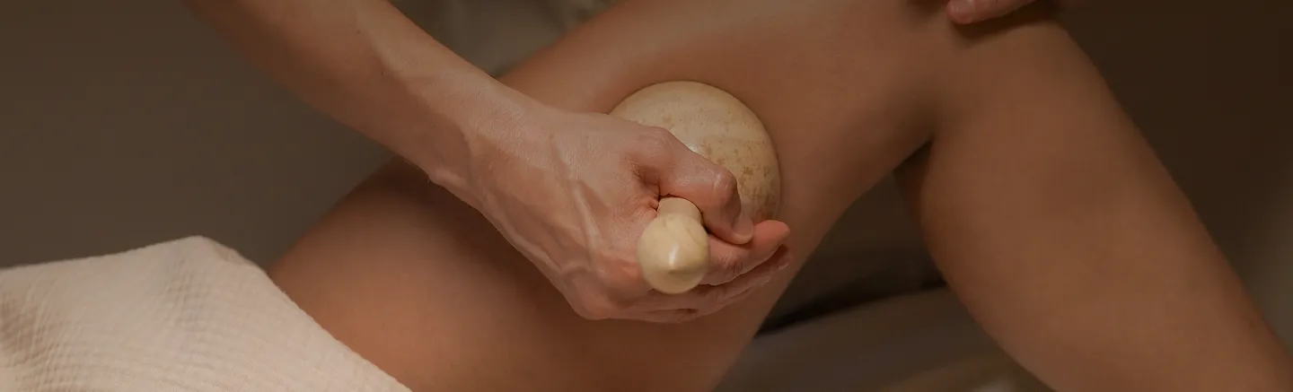 Una mano sostiene una herramienta de masaje de madera y realiza un masaje anticelulítico en un muslo.