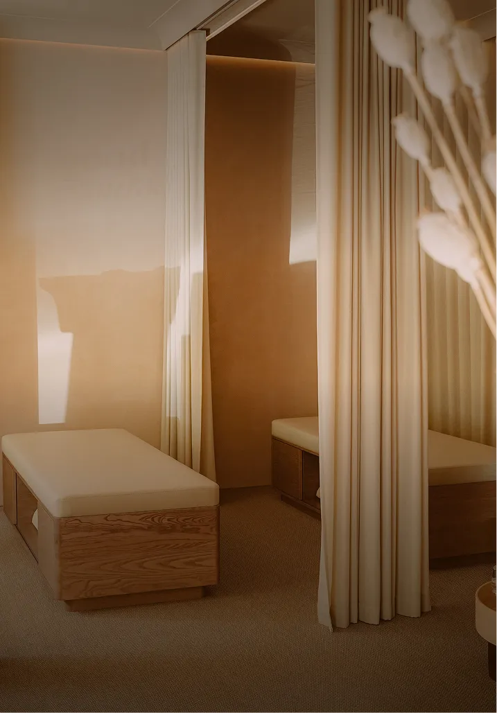 Sala de tratamiento con cortinas claras, cama de madera y flores secas en una maceta.