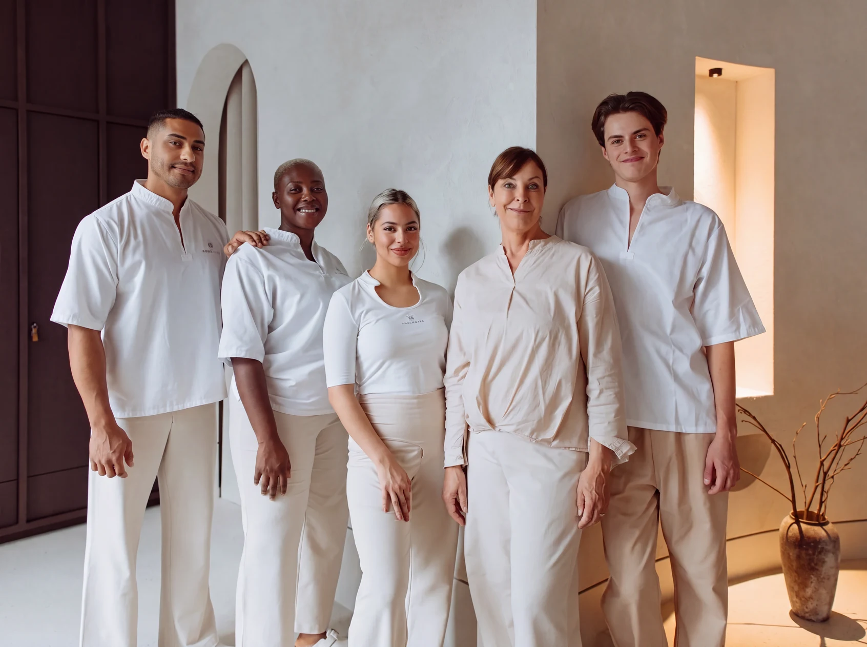 Foto de grupo de 5 Experts en masaje con uniformes blancos y beige con el logo de Pluma, posando en una sala luminosa con un jarrón al fondo.