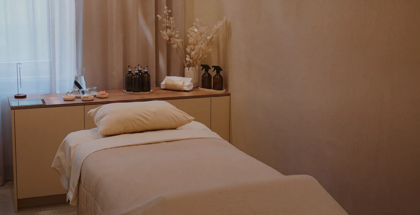 Sala de tratamiento tranquila con camilla y cojín, una mesa con aceites de aromaterapia, toallas y flores secas en un jarrón.