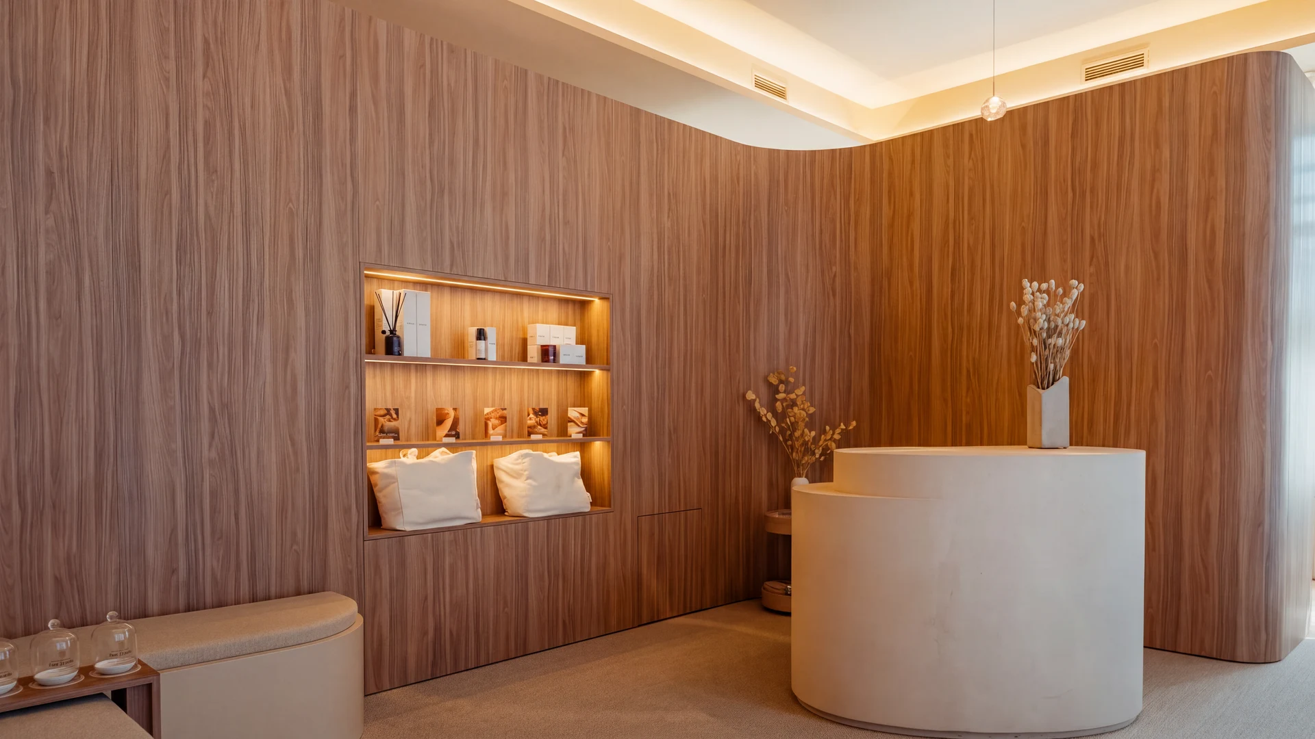 Zona de recepción de un Studio de Pluma con un mostrador redondo blanco, pared de madera con estantería iluminada y dos cojines con la inscripción 'be good to yourself'.
