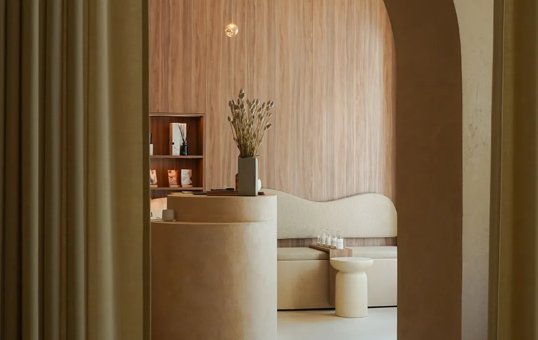 Zona de espera minimalista en un Studio de Pluma con mostrador de recepción redondo, banco con respaldo ondulado y revestimiento de madera en tonos neutros.